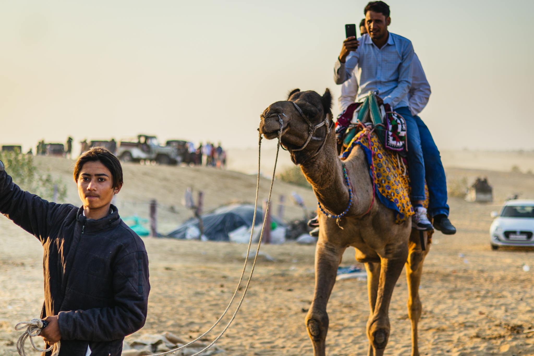 A man rides a camel in the desert, taking photos, under a clear sky.